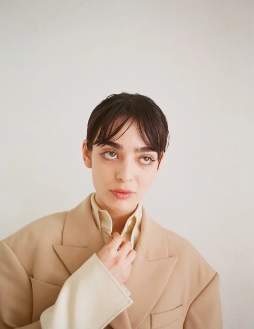 Portrait of a brunette adult female with eyes looking up, wearing a beige blazer and a cream-colored collared shirt in a photo studio.