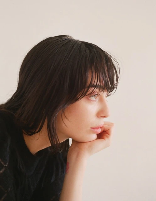 Close-up studio portrait of a young adult woman with black hair resting her chin on her hand against a plain, off-white background.