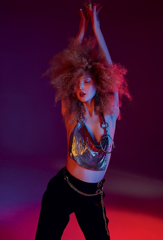 Young woman with afro hairstyle poses in a metallic top and black pants against a vibrant pink and purple studio backdrop.