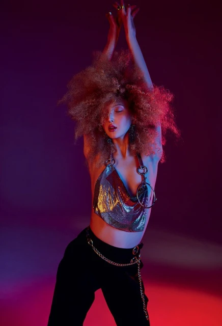 Young woman with afro hairstyle poses in a metallic top and black pants against a vibrant pink and purple studio backdrop.