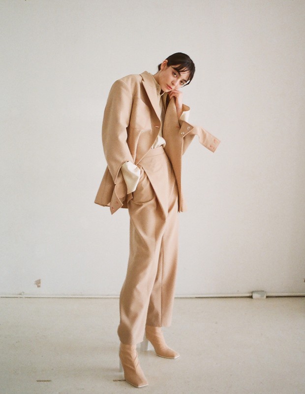 Full-length shot of a woman in a tan suit and matching boots, posing in a minimalist studio with neutral colors and modern fashion.