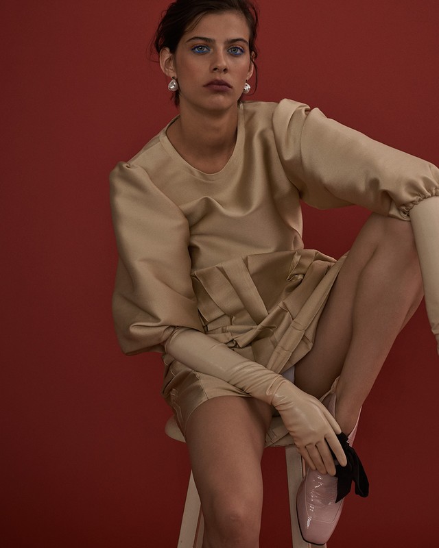 Woman in a light-brown dress and gloves poses while seated on a stool against a reddish-brown background in a fashion shot.