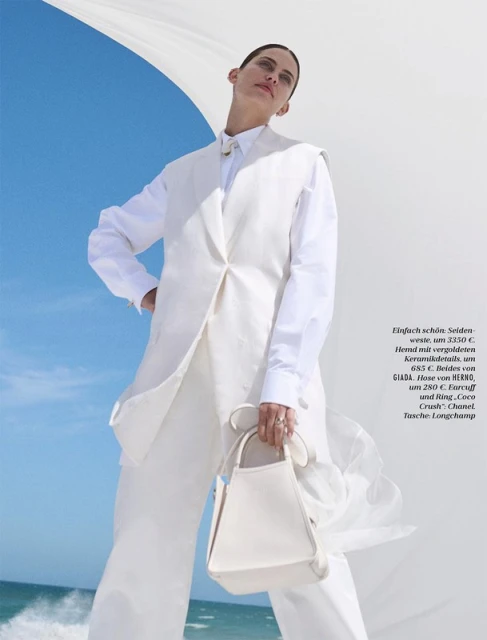 A woman models white vest, pants, shirt, and handbag on a beach. The blue sky meets white fabric in the background.