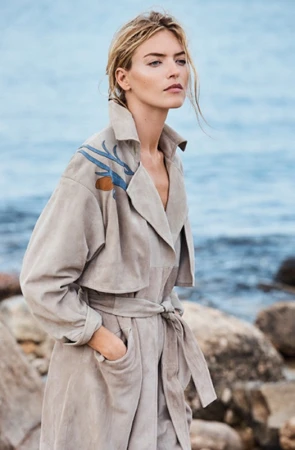 Blonde woman wearing a beige belted trench coat with blue painted shoulder details by the rocky coast.