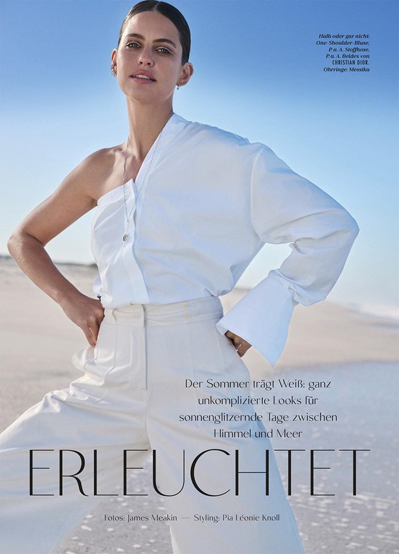 Model in white one-shoulder blouse and trousers posing on a sandy beach under clear sky