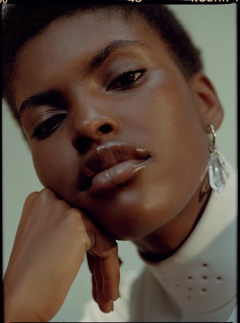 Close-up portrait of a dark-skinned woman with short hair, glossy lips, and a white collared shirt with decorative cutouts, wearing large crystal earrings.