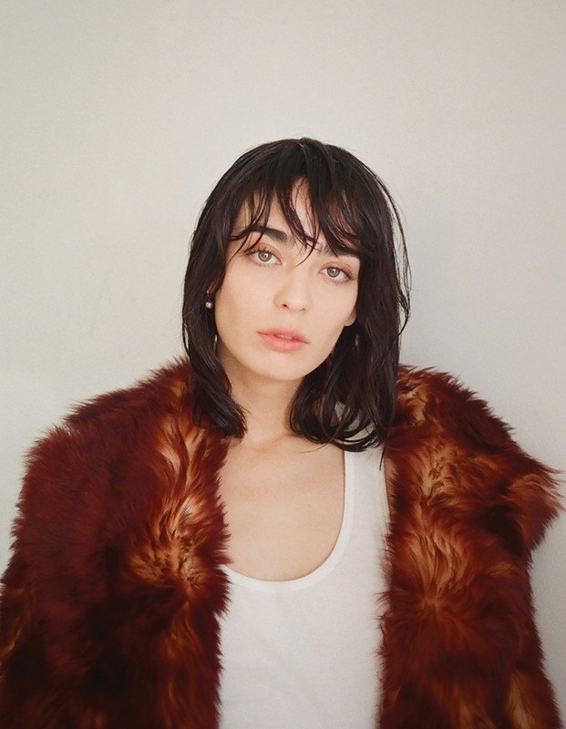 Woman with dark wet-look hair wearing a white tank top and a brown faux fur coat against a plain wall.