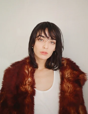Woman with dark wet-look hair wearing a white tank top and a brown faux fur coat against a plain wall.