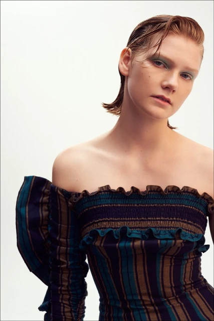 Striking portrait of a fair-skinned young woman with slicked-back auburn hair, wearing a form-fitting, off-the-shoulder striped dress.