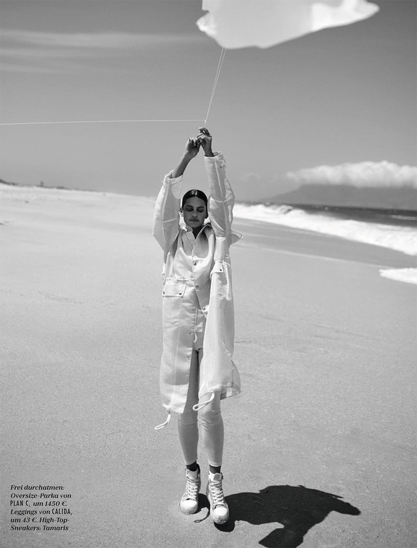 A woman in a light-colored outfit stands on a beach holding a cloud-shaped balloon in a black and white fashion photography shot.
