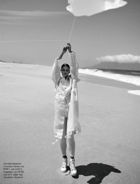 A woman in a light-colored outfit stands on a beach holding a cloud-shaped balloon in a black and white fashion photography shot.