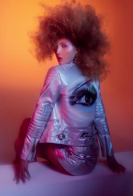 Adult female model with afro, wearing a silver jacket with an eye painted on the back, and matching miniskirt, looking over shoulder.