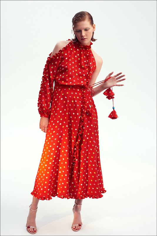 Adult woman in a red dress with white polka dots, one shoulder exposed, ruffled trim, and tan heels posing in a photo studio.