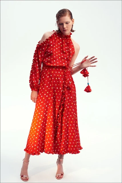 Adult woman in a red dress with white polka dots, one shoulder exposed, ruffled trim, and tan heels posing in a photo studio.