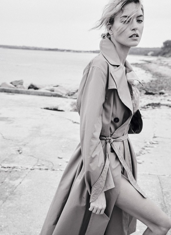 A woman poses in a trench coat on a concrete walkway near a body of water under an overcast sky in a monochrome photograph.