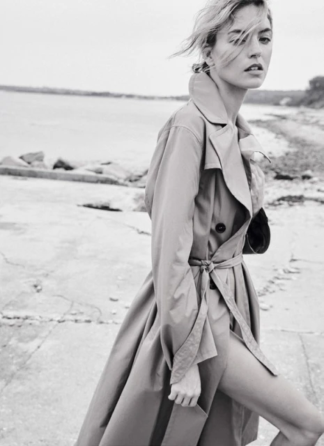 A woman poses in a trench coat on a concrete walkway near a body of water under an overcast sky in a monochrome photograph.