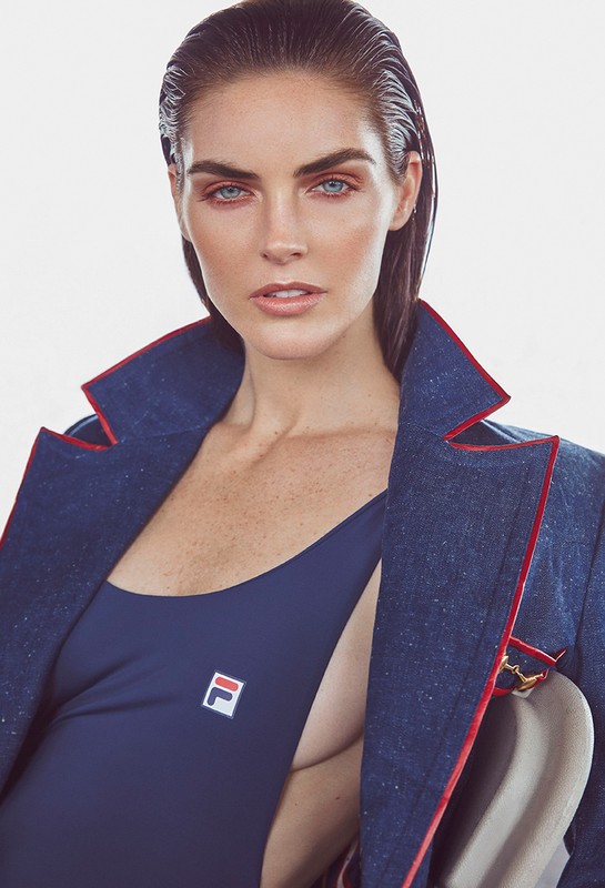 Portrait of a woman with slicked-back hair, wearing a dark blue swimsuit and blazer with red trim against a white background.