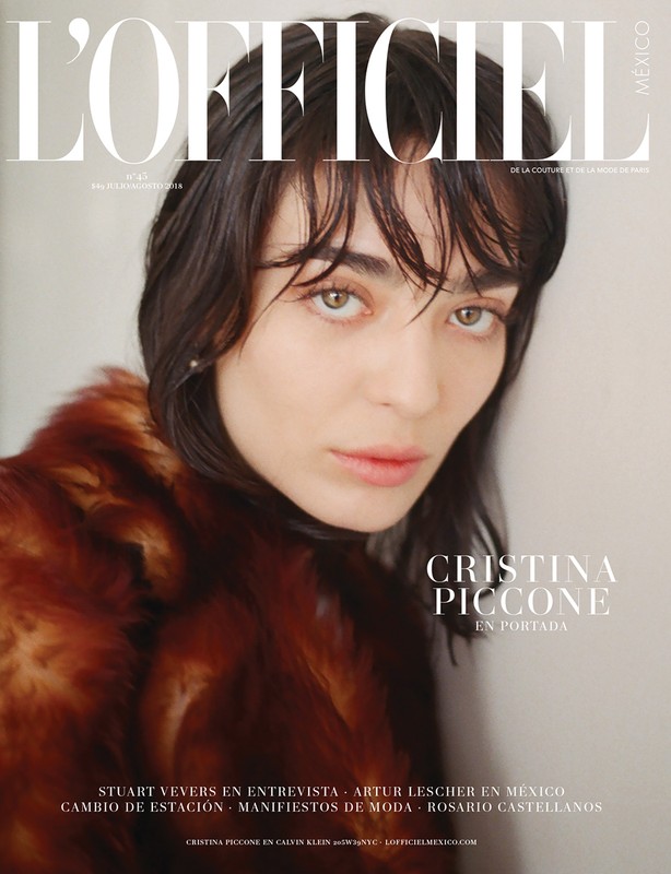 Magazine cover portrait of a dark-haired woman wearing a rust-colored fur coat against a plain wall.