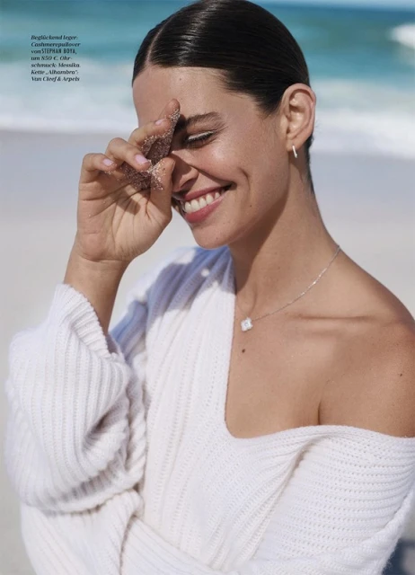 Smiling woman poses on beach with sand in hand. She's wearing a white sweater and silver necklace with an ocean backdrop.