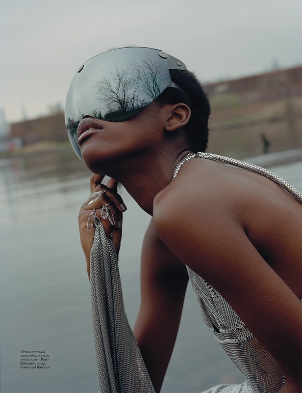 Young Black female model poses in a futuristic mirrored visor and silver chainmail dress next to a river on an overcast day.