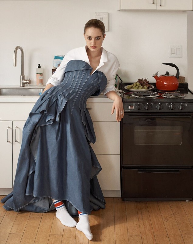 Woman in a strapless denim dress and white shirt posing in a kitchen with stove, kettle, and fruit, creating a fashion editorial image.