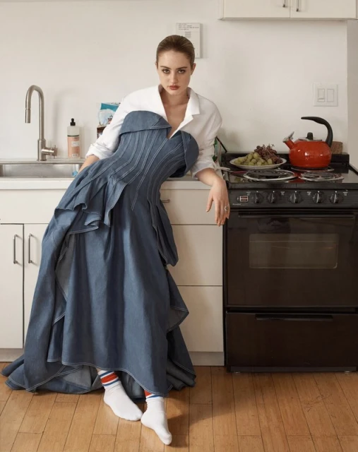 Woman in a strapless denim dress and white shirt posing in a kitchen with stove, kettle, and fruit, creating a fashion editorial image.