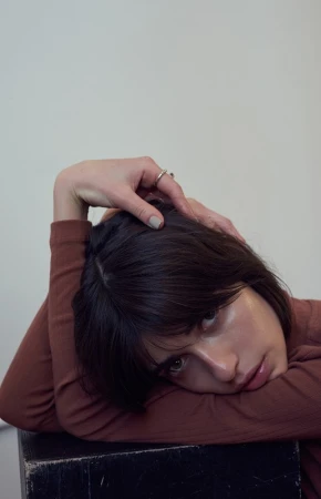 Close-up portrait of a woman with dark hair resting her head on her arms against a plain white background.