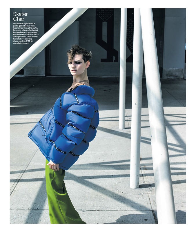 Fashion shot of a model wearing a large blue puffer jacket and green pants, posing outdoors against a backdrop of white poles.