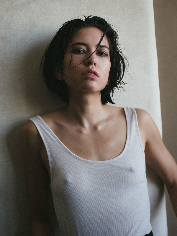Portrait of a young adult woman with short dark hair, wearing a white tank top, against a neutral beige background in a studio setting.