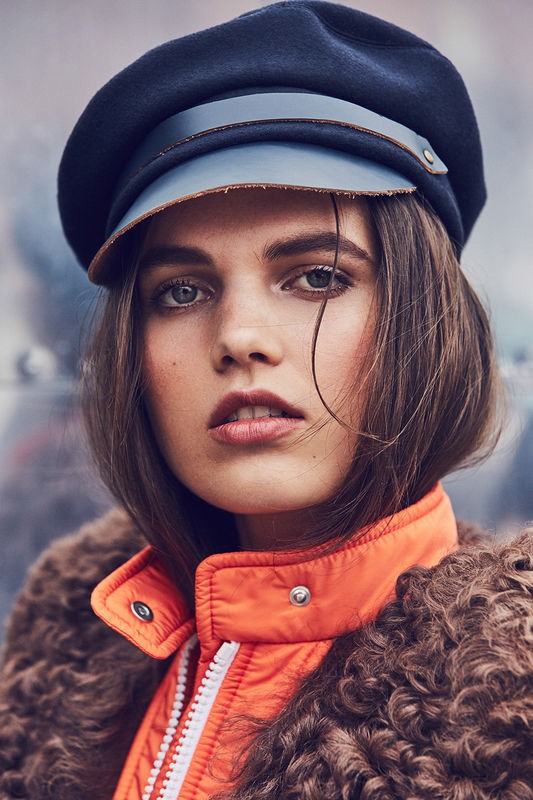 Portrait of a young woman with brown hair wearing a blue cap and orange coat with fur collar against a blurred background.