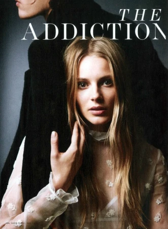 Close-up fashion shot: A young woman with blonde hair, wearing a white lace-detailed top, poses in a studio with another person behind.