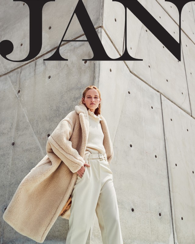 Fashion editorial shot of an adult woman wearing a neutral-toned teddy coat, sweater, and pants standing against a geometric concrete wall.