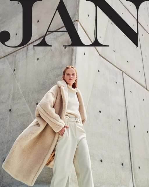 Fashion editorial shot of an adult woman wearing a neutral-toned teddy coat, sweater, and pants standing against a geometric concrete wall.