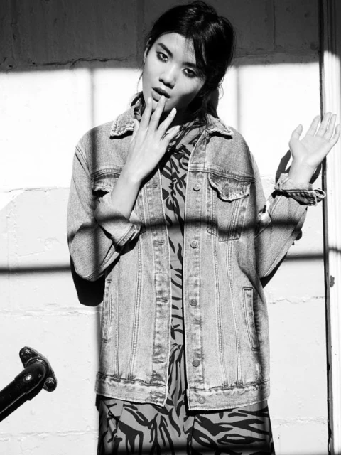 Black and white portrait of an Asian woman in a denim jacket and patterned dress, posing near a window casting shadows on the wall.