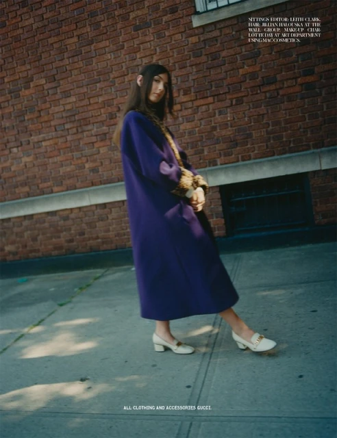 A fashion shot capturing a model in a long purple coat and ivory shoes, posing against a brick wall and sidewalk, showcasing street style.