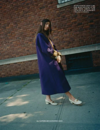 A fashion shot capturing a model in a long purple coat and ivory shoes, posing against a brick wall and sidewalk, showcasing street style.