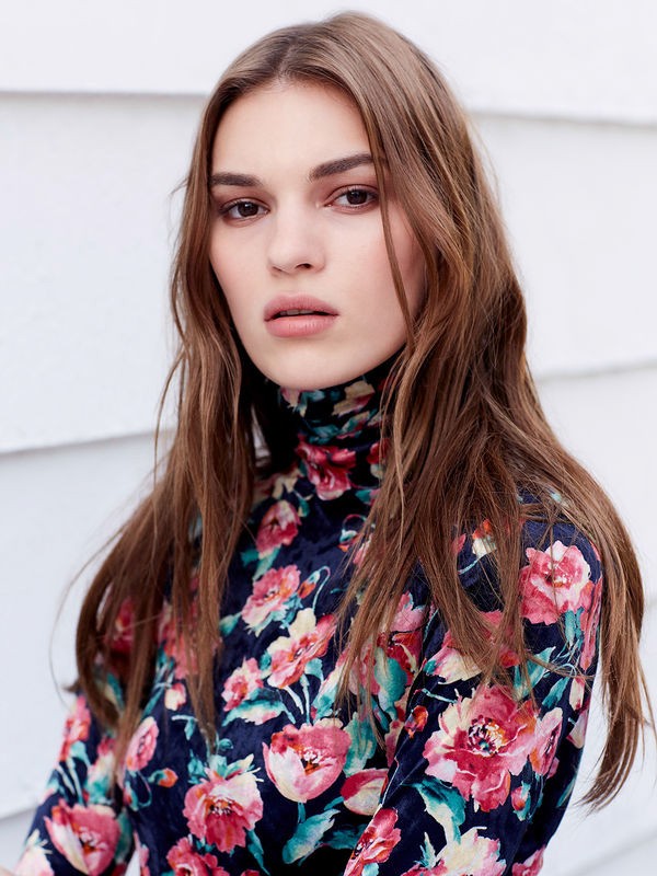 Portrait of a young adult female with long brown hair, wearing a floral pattern top against a white shiplap background.