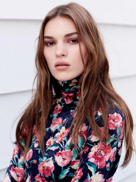 Portrait of a young adult female with long brown hair, wearing a floral pattern top against a white shiplap background.