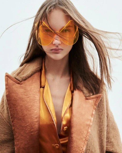 A fashionable woman with flowing brown hair poses in a coat with orange sunglasses and a satin shirt in a fashion style portrait.