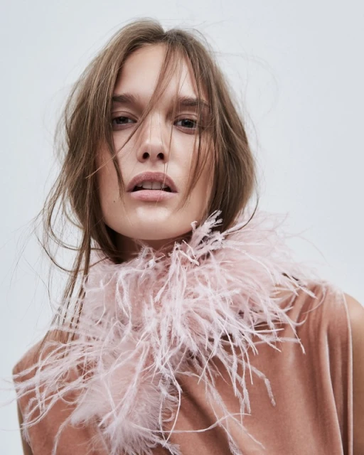 Close-up portrait of a fair-skinned young woman with light brown hair wearing a blush-colored velvet top and a feathered collar against white.