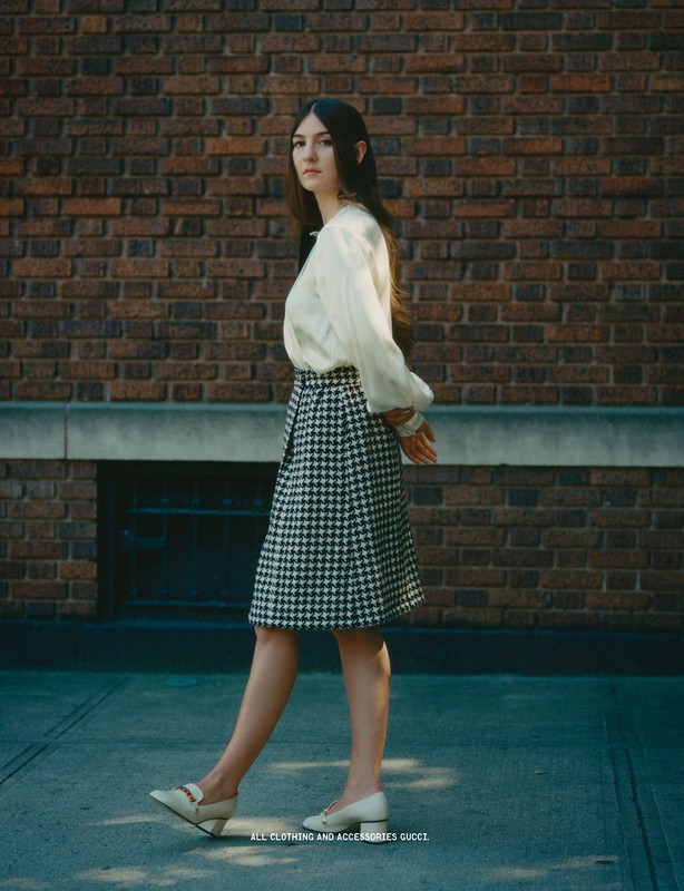 Full shot of a woman in a white blouse and patterned skirt walking past a brick wall on a city street, looking at the camera.