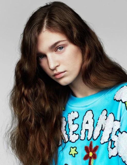 A young woman with long brown hair poses in a blue sweatshirt with colorful embroidered accents against a neutral background.
