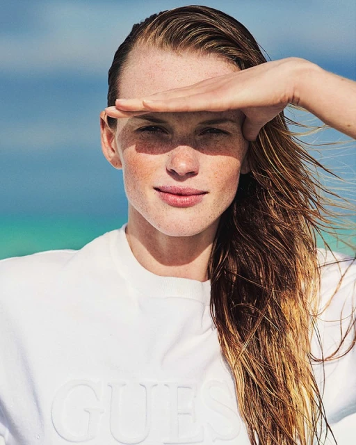 Portrait of a woman with long, wet hair shading her eyes with her hand in front of an aqua ocean wearing a white "GUESS" sweatshirt.