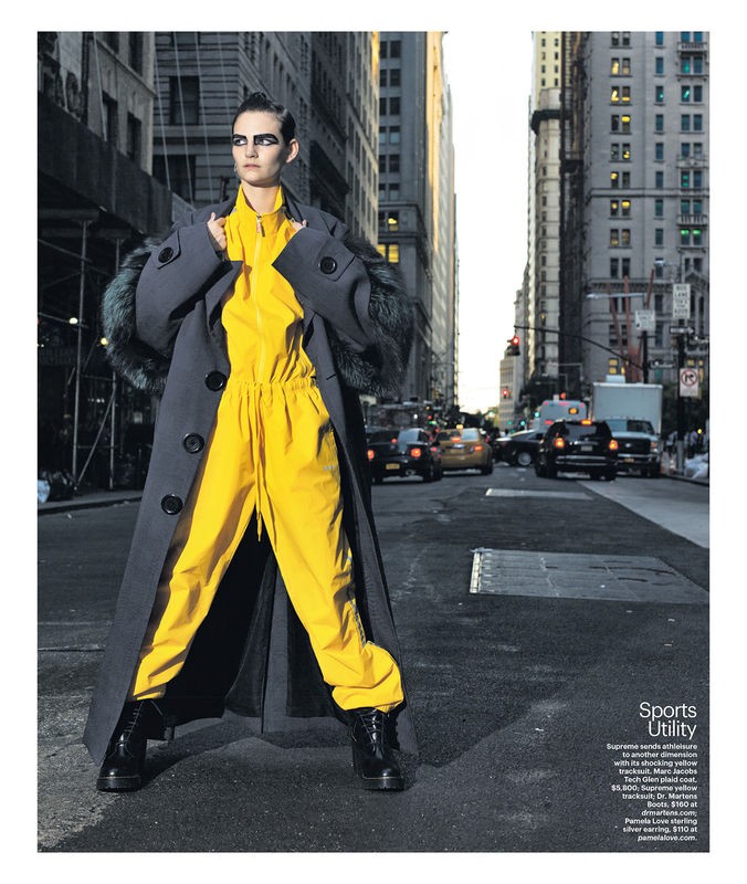 Fashion shot of a stylish adult female in a bright yellow jumpsuit and oversized grey coat posing on a city street with buildings and cars.
