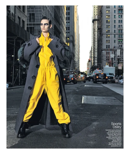 Fashion shot of a stylish adult female in a bright yellow jumpsuit and oversized grey coat posing on a city street with buildings and cars.