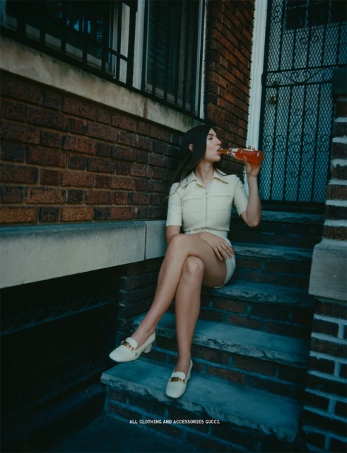 Brunette woman in a cream-colored outfit sits on outdoor stairs drinking a beverage, set against a brick wall and a blue door.