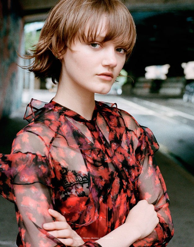 Fashion shot of a young woman with brown hair in a red and black patterned dress. She poses with arms crossed in an urban tunnel.