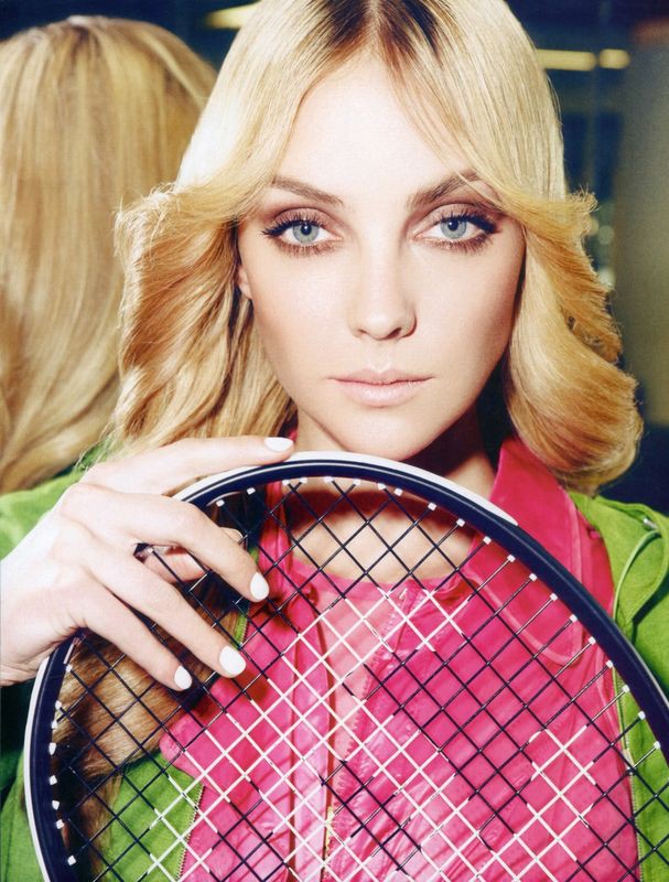 Woman with blonde hair and blue eyes poses with a tennis racket, wearing a green jacket and pink shirt in an indoor setting.