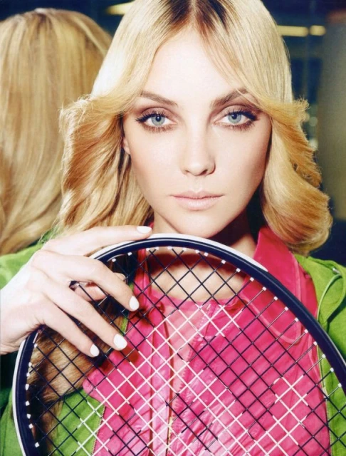 Woman with blonde hair and blue eyes poses with a tennis racket, wearing a green jacket and pink shirt in an indoor setting.