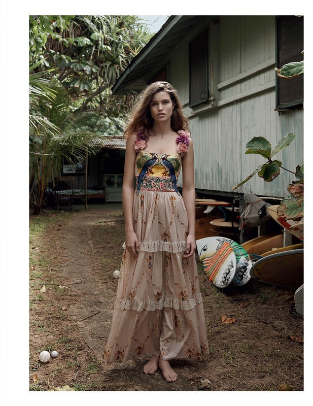 A woman in a floral dress stands barefoot on a dirt path near a white house surrounded by tropical greenery in an outdoor setting.